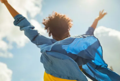 Rear view of a joyful person with curly hair and a flowing blue shirt raising their arms toward the sun