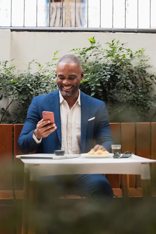 Man in a blue suit sitting at an outdoor café table, smiling at his phone with a coffee and pastry beside him