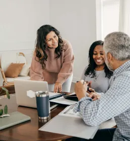 Three colleagues having a friendly discussion around a table with laptops and notebooks in a cozy, modern office
