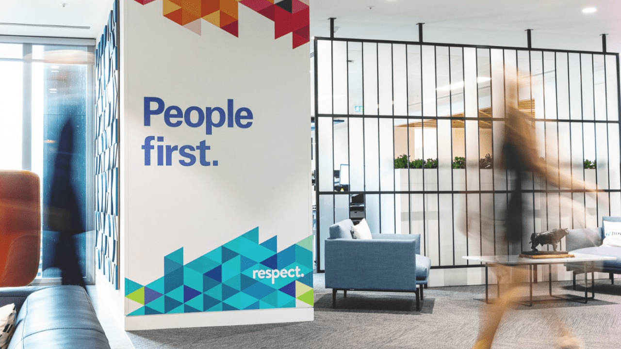 A view inside our London office, of a female staff member walking past a wall with People First printed on it