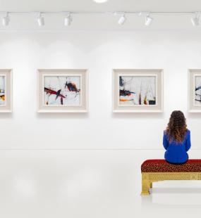 A young woman sits on a bench in a museum admiring the modern art on the wall in front of her
