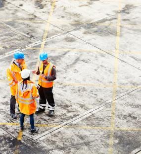 Three construction workers in hard hats and high vis jackets stand talking on a building site