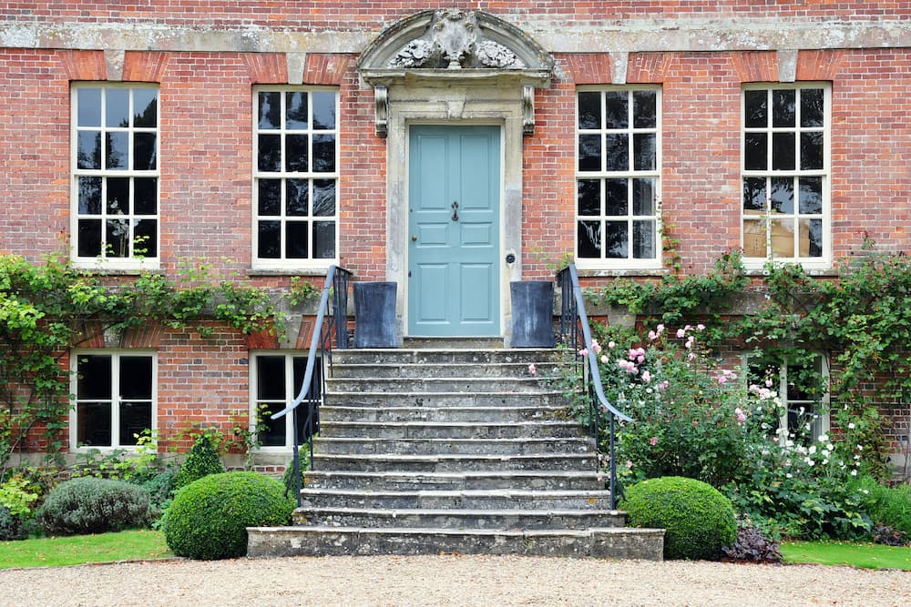 The front of a red brick country house, with large windows and a pale blue door at the top of a set of stone steps leading into a manicured garden