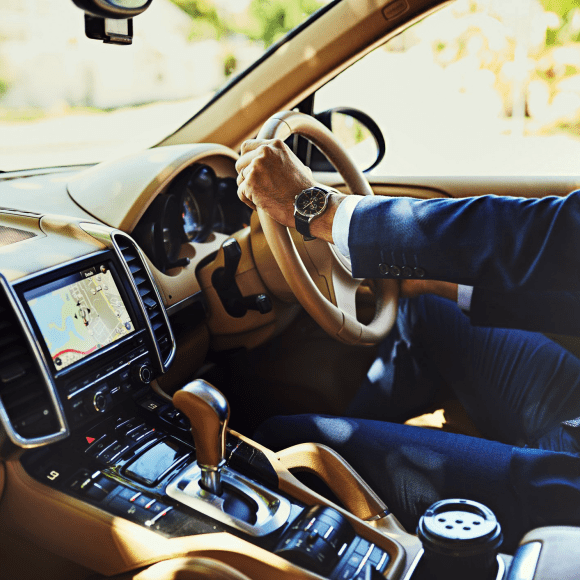A handsome man in a tailored suit sits in an expensive car with his hands on the steering wheel