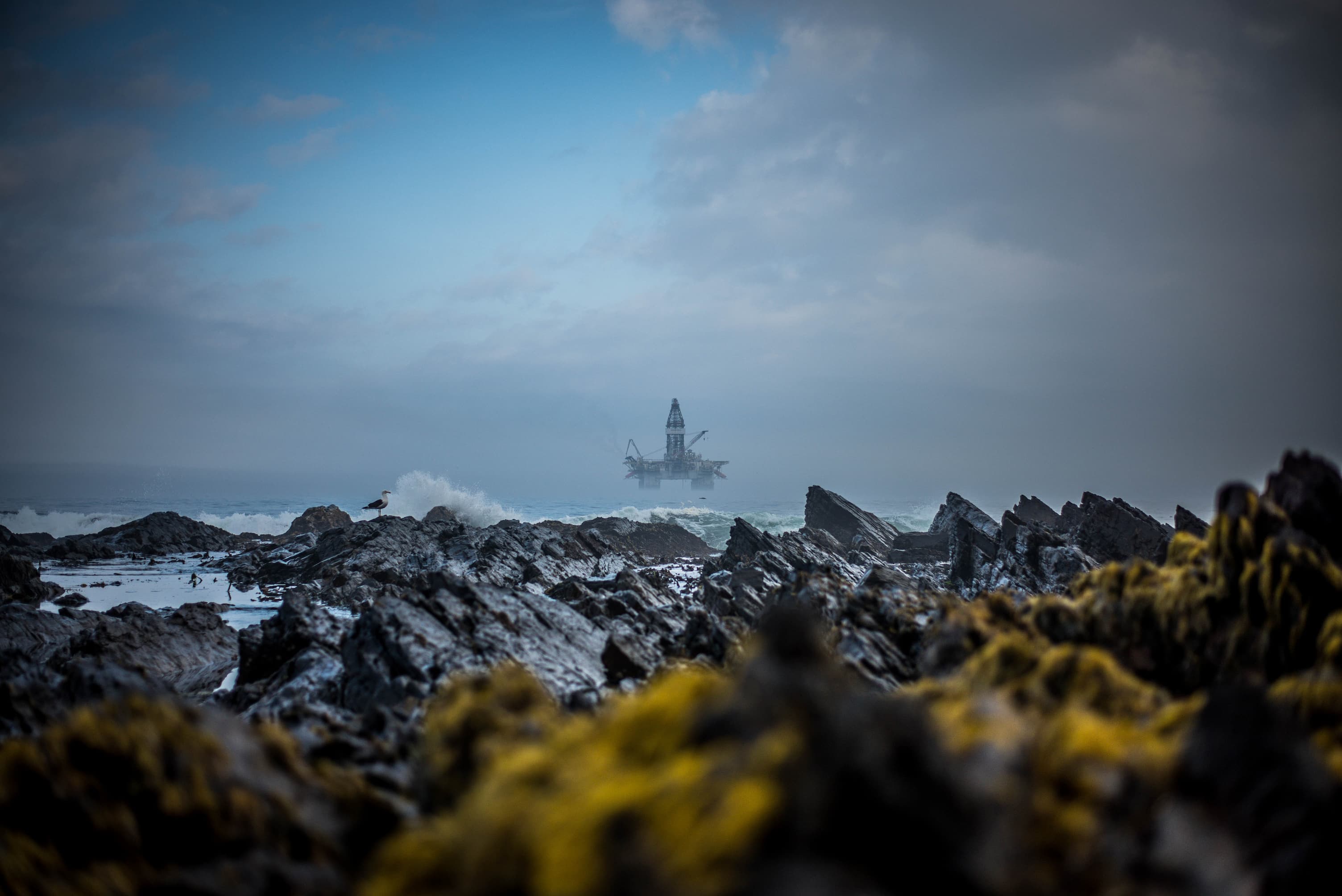 Oil rig in distance out to sea, crashing waves against rocks in foreground