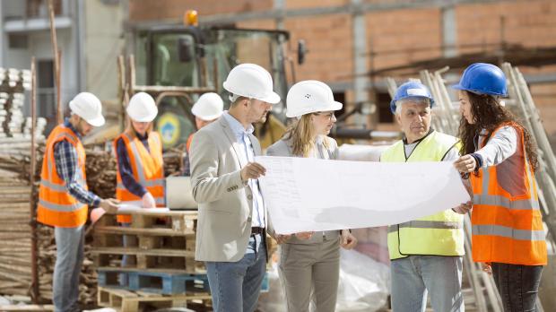 A group of builders, architects, and surveyors stand on a building site consult a paper plan 