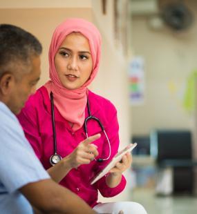 A female doctor in a pink headscarf and pink scrubs speaks with a patient in the hallway of a hospital