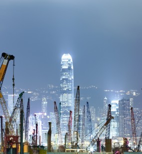 A nighttime view of the Hong Kong skyline with dozens of construction cranes in the foreground and illuminated skyscrapers behind