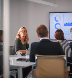  blonde business woman speaks to a colleague during a board meeting, where everyone around the table sits looking at a presentation on a large screen