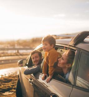 A young family take a day trip to the beach. They sit in their car looking out of the windows at the sea. The dad sits in the driver's seat, while his wife and son sit in the back seat. The little boy leans out of the window to get a better view.