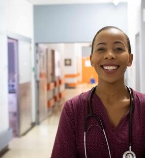 A nurse in red scrubs and a stethoscope stands in a hospital corridor and smiles at the camera