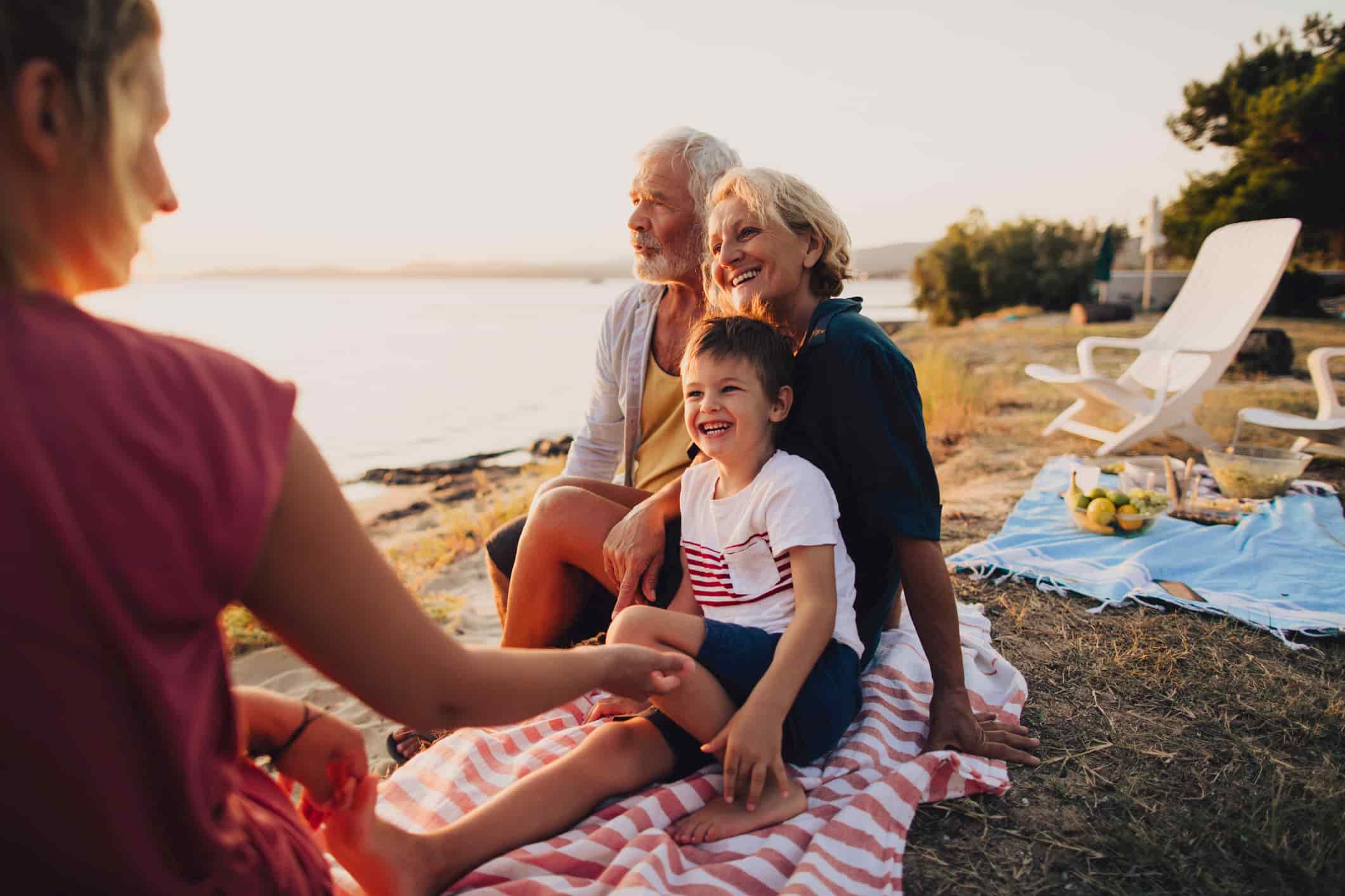 An attractive older couple sits on a picnic blanket by the sea with their daughter and grandson. They are all smiling, and the man looks out at the sea
