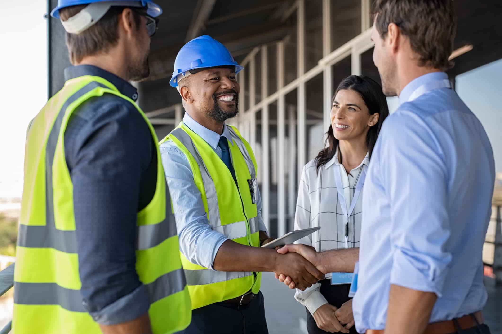A team of four engineers and architects speak on a construction site. Two wear high visibility jackets and safety hats. One smiling man in a high vis shakes the hand of a man in a blue shirt