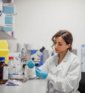 A female researcher or biochemist, sitting in a lab wearing a lab coat and blue plastic gloves, analyses a specimen in a test tube