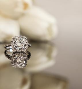 A diamond ring sitting on a reflecting surface in front of some white roses