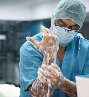 A doctor in scrubs and a face mask washes his hands and wrists in the operating room