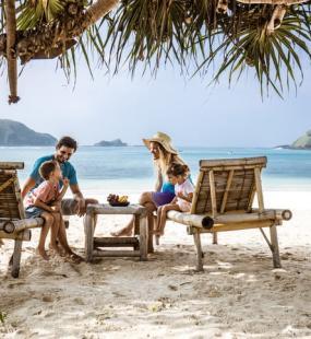 A happy young family talk while relaxing on deck chairs on a beautiful sandy beach below palm trees