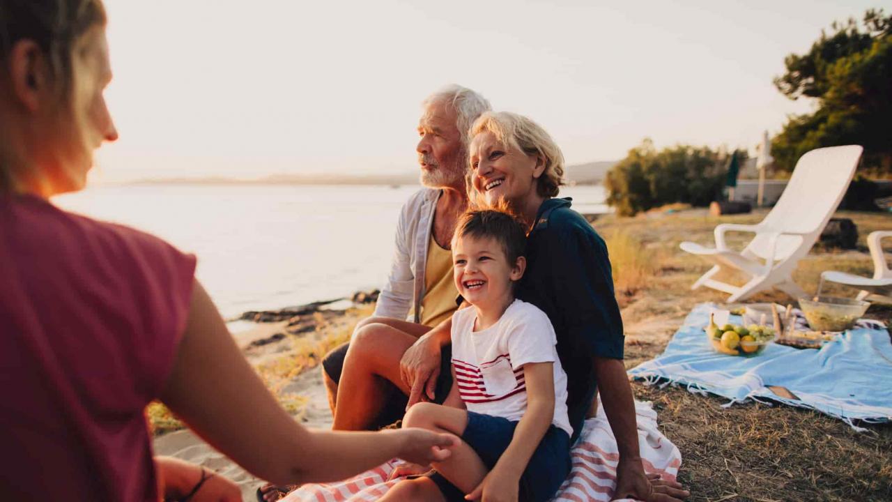 An attractive older couple sits on a picnic blanket by the sea with their daughter and grandson. They are all smiling, and the man looks out at the sea