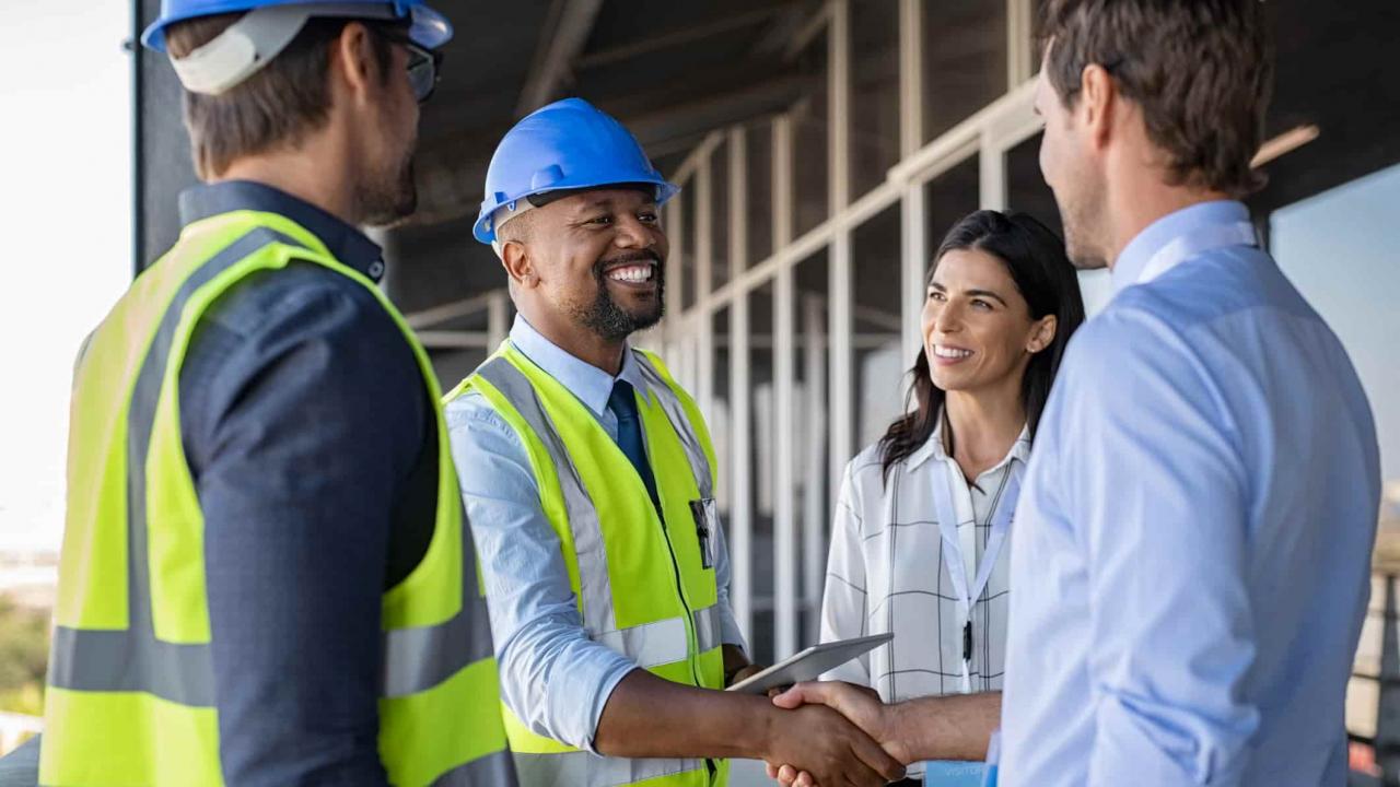 A team of four engineers and architects speak on a construction site. Two wear high visibility jackets and safety hats. One smiling man in a high vis shakes the hand of a man in a blue shirt