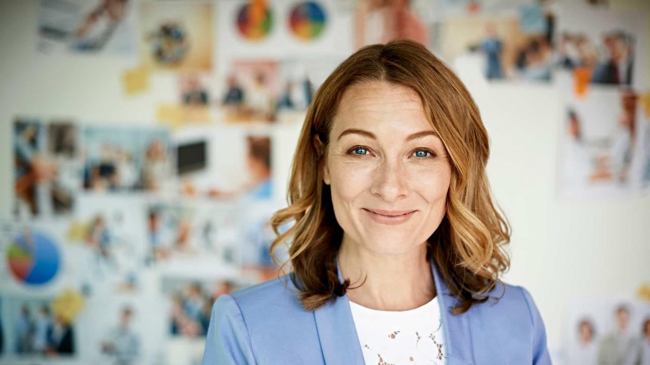 A portrait of a smiling corporate businesswoman in a suit, standing in front of a wall of photographs