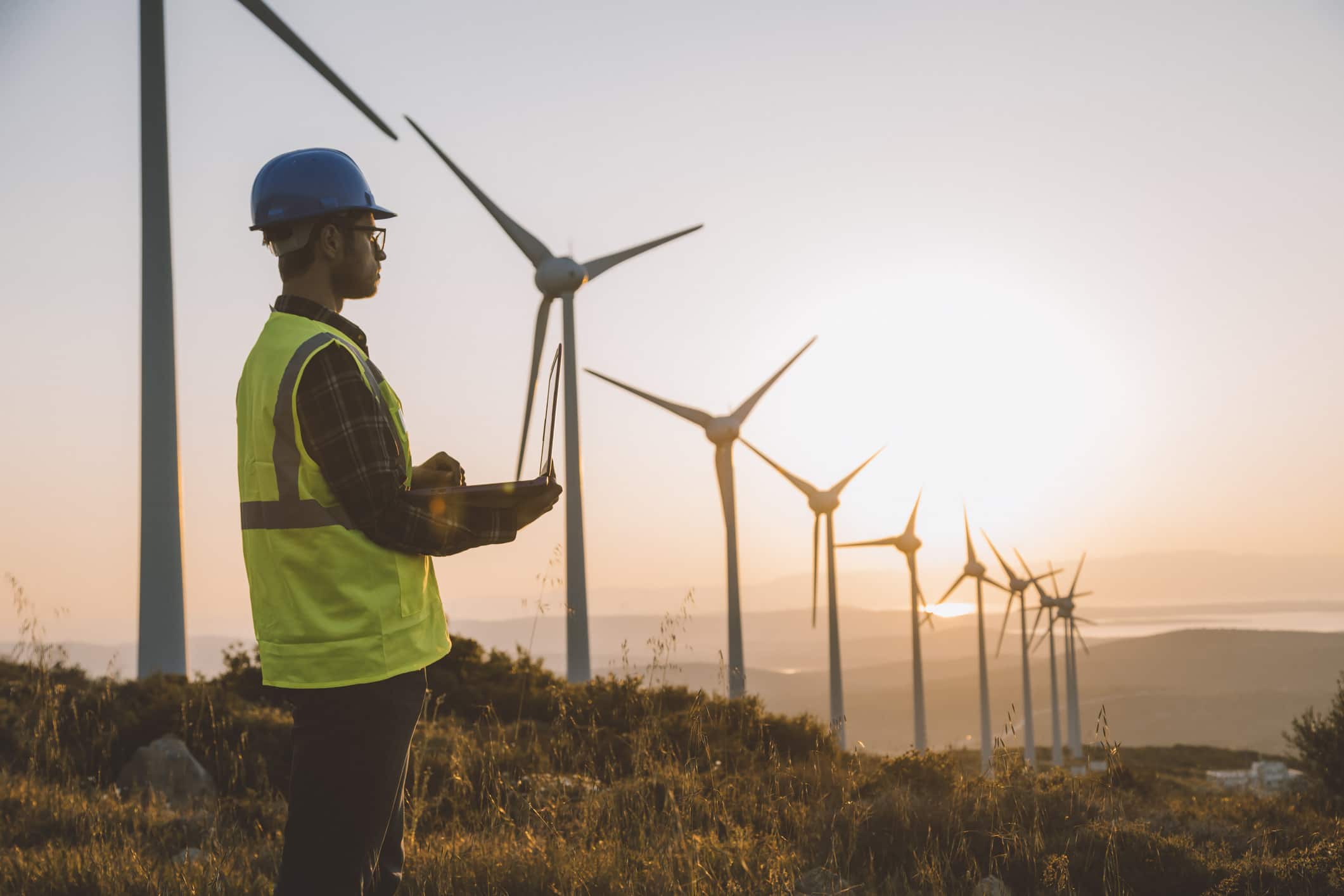 An engineer in high vis and hard hat stands looking across a field of wind turbines