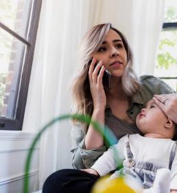 A mother holds her baby and tests her temperature while on the phone 