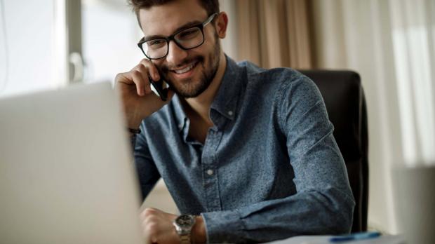 A smiling young businessman sits at his computer and speaks on the phone