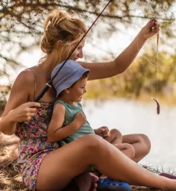 A woman and her young son are fishing by the side of a sunny lake. She holds up a small fish on the line as he smiles happily