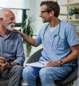 A young male doctor sits next to an older patient. The patient is speaking to the doctor, who is smiling and has his right hand on the patient's left shoulder