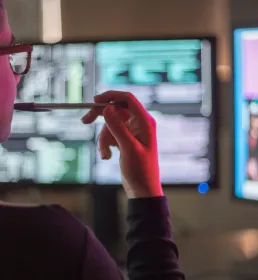 A computer technician sits looking at two monitor screens. She is illuminated by the red light from her screens