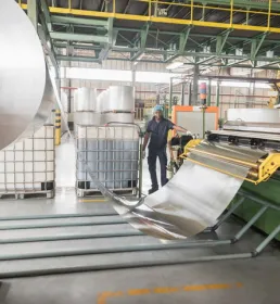 A male factory worker stands in a warehouse by a large roll of aluminium