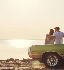 A young couple lean on the bonnet of a vintage green car and watch the sun set over the sea