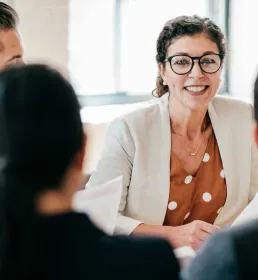 A woman in a business suit sits at a table with her colleagues and smiles as they talk