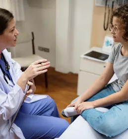 An female patient sits on a bed talking with her doctor, who is holding her chart