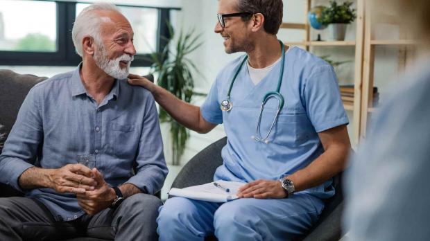 A young male doctor sits next to an older patient. The patient is speaking to the doctor, who is smiling and has his right hand on the patient's left shoulder