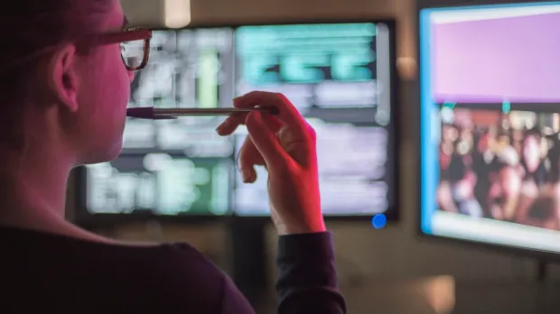A computer technician sits looking at two monitor screens. She is illuminated by the red light from her screens