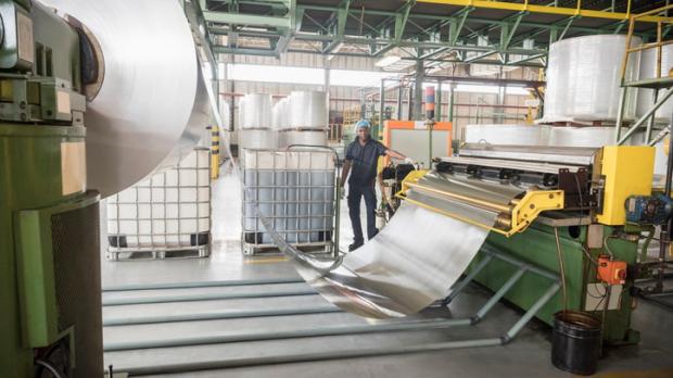 A male factory worker stands in a warehouse by a large roll of aluminium 