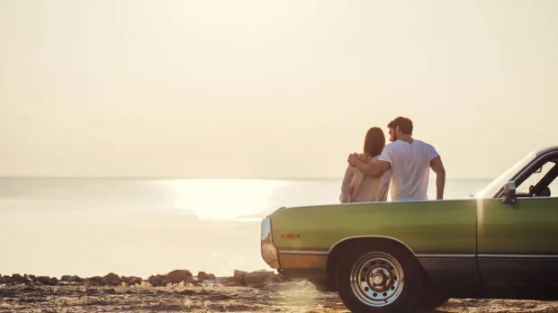 A young couple lean on the bonnet of a vintage green car and watch the sun set over the sea