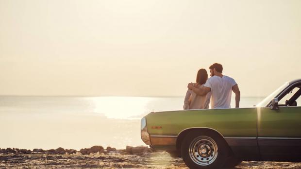A young couple lean on the bonnet of a vintage green car and watch the sun set over the sea