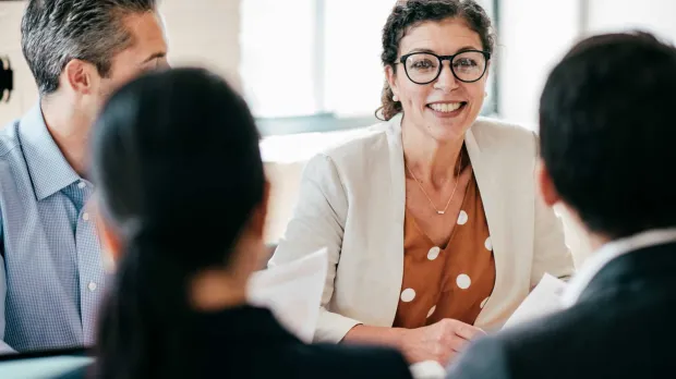 A woman in a business suit sits at a table with her colleagues and smiles as they talk