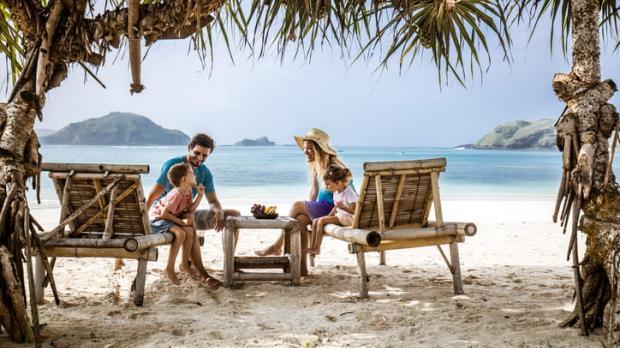 A happy young family talk while relaxing on deck chairs on a beautiful sandy beach below palm trees