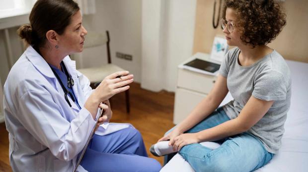 An female patient sits on a bed talking with her doctor, who is holding her chart