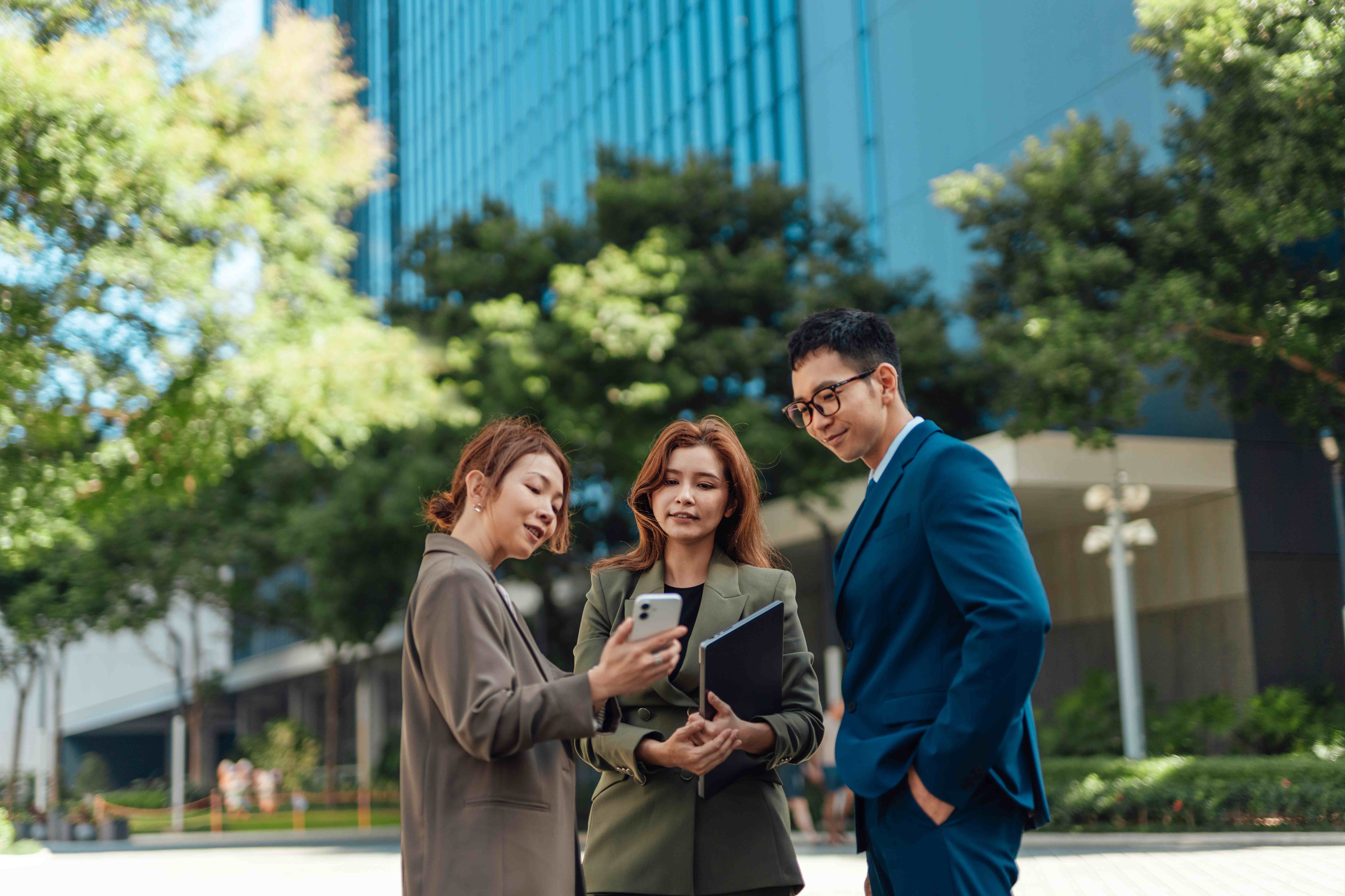 Asian business people taking a break from work, sharing a smartphone, talking and smiling, standing in business park. Teamwork and collaboration. Employee engagement and wellbeing.