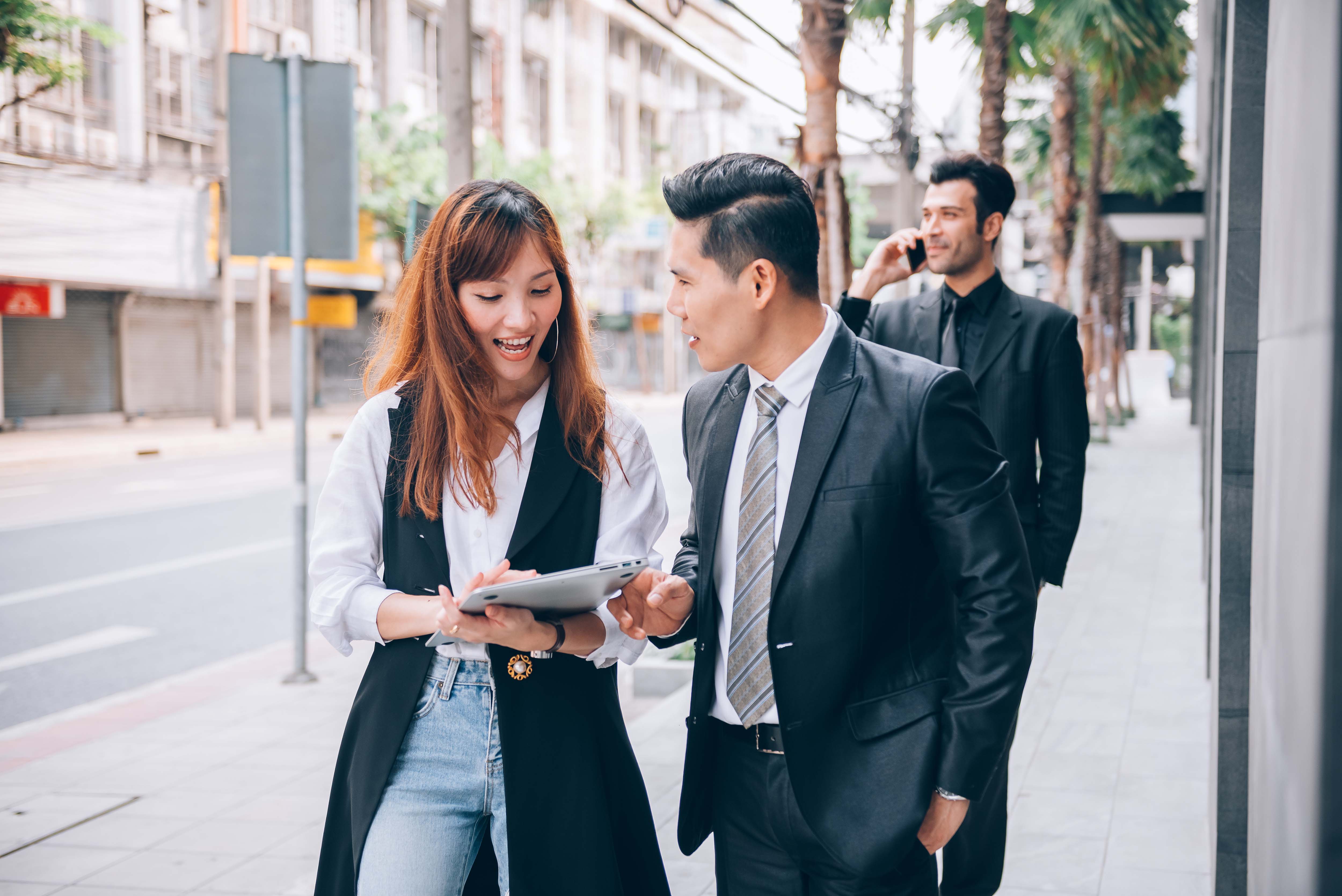 Group of Asian business professionals walking and discussing work outdoors in a city business district, one woman holding a tablet while talking to a colleague in a suit.