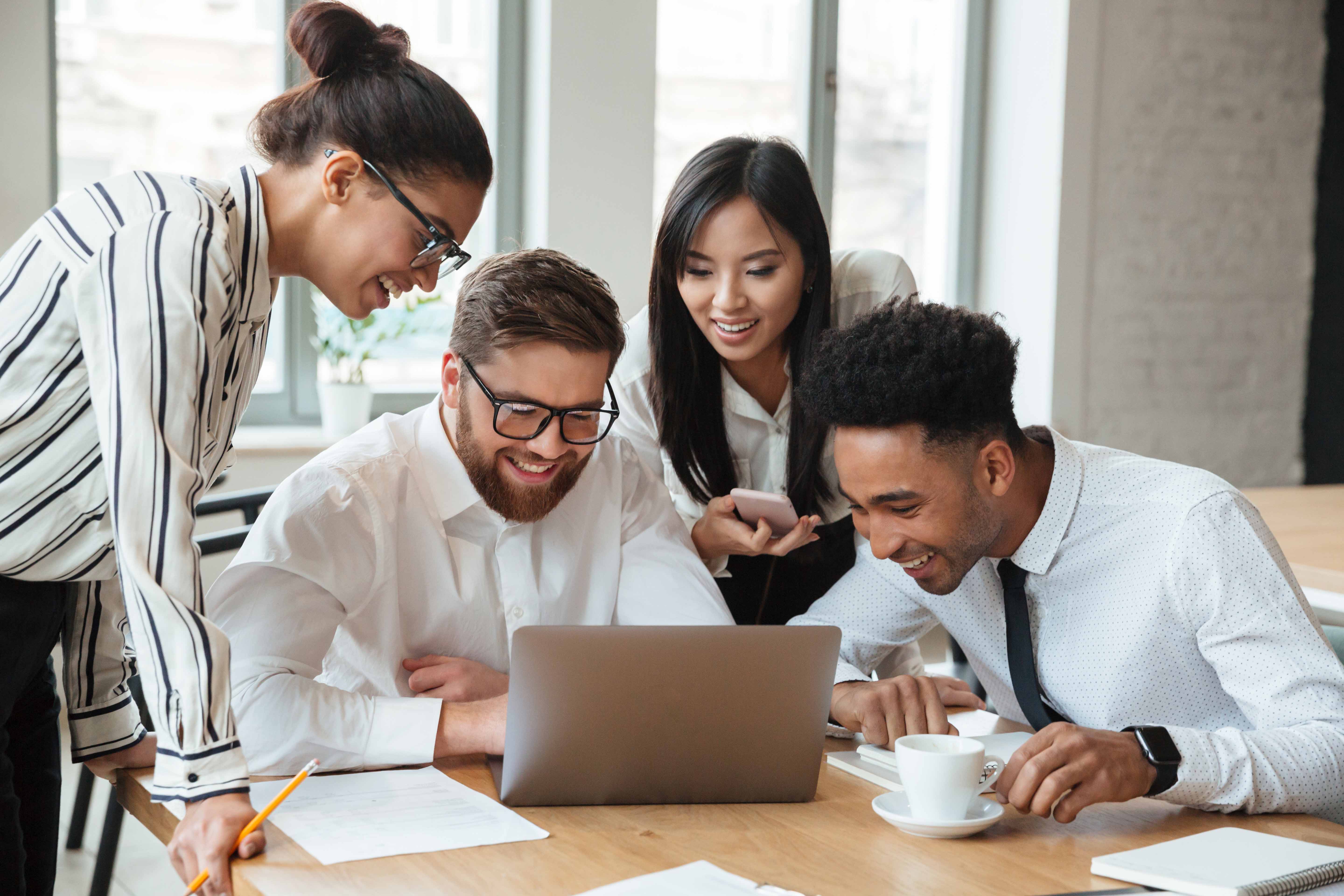 A diverse group of young professionals gathered around a laptop, smiling and collaborating in a modern office setting.