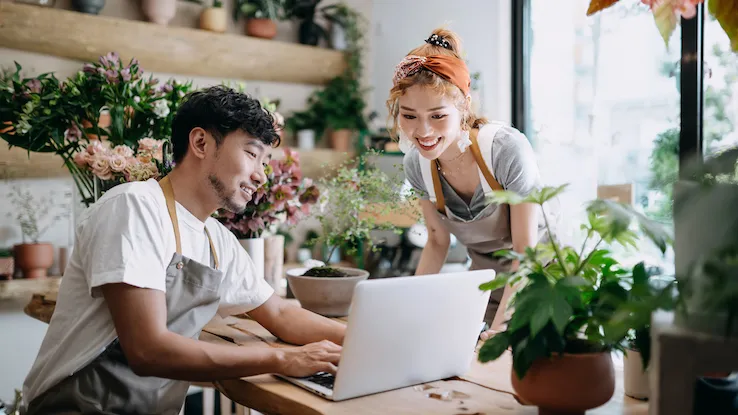Smiling young Asian couple, the owners of small business flower shop, discussing over laptop on counter against flowers and plants. Start-up business, business partnership and teamwork. Working together for successful business