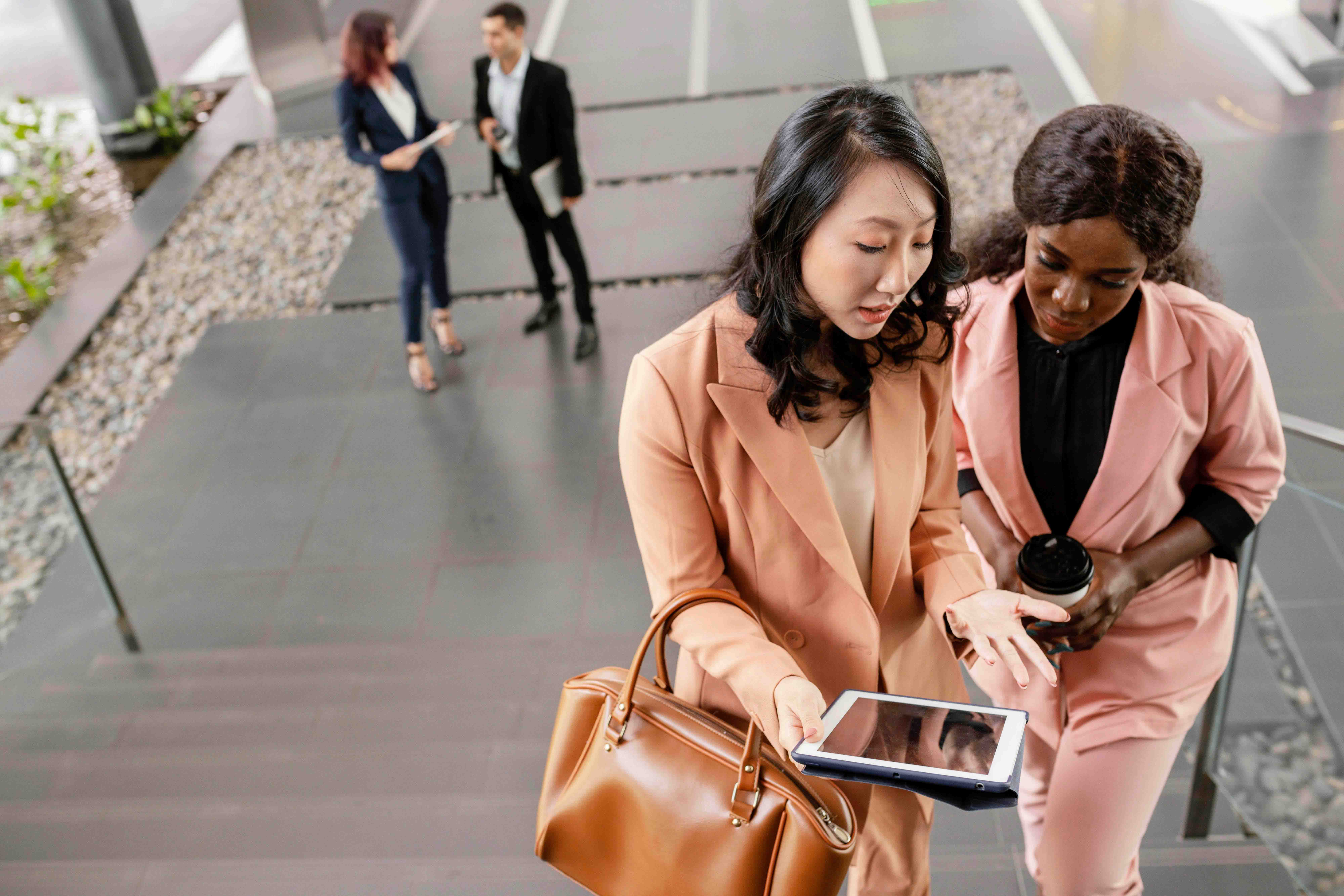 Two professional women in conversation while walking up stairs, one holding a tablet and the other a coffee cup, with colleagues in the background.