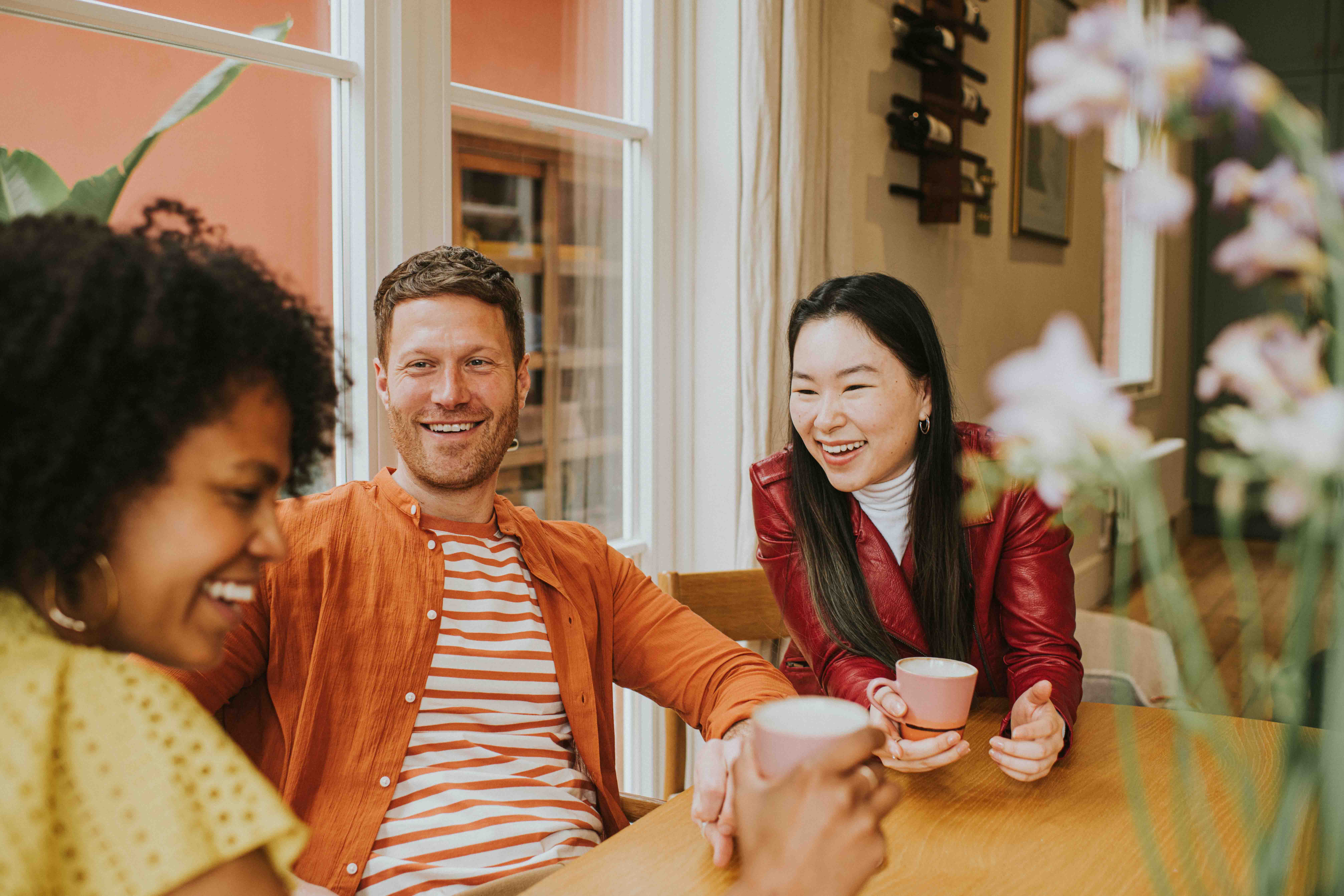 Three colleagues sharing a laugh and enjoying coffee together around a wooden table, capturing a moment of authentic connection and team culture