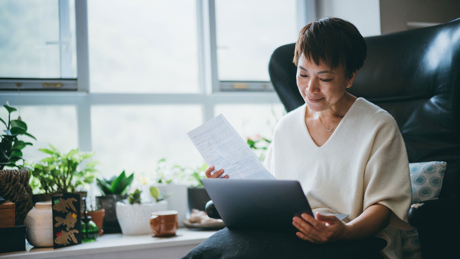 A middle-aged woman sitting comfortably in a bright, plant-filled room, smiling as she looks at her laptop and holds a printed document in her hand