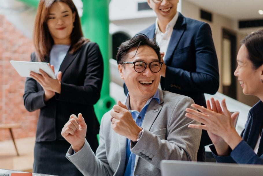 Smiling business professionals celebrating a success in the office, with one man laughing and colleagues clapping around him.
